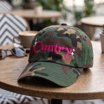Country Word Art embroidered hat in Green Camo resting on a flat surface, lifestyle image 2