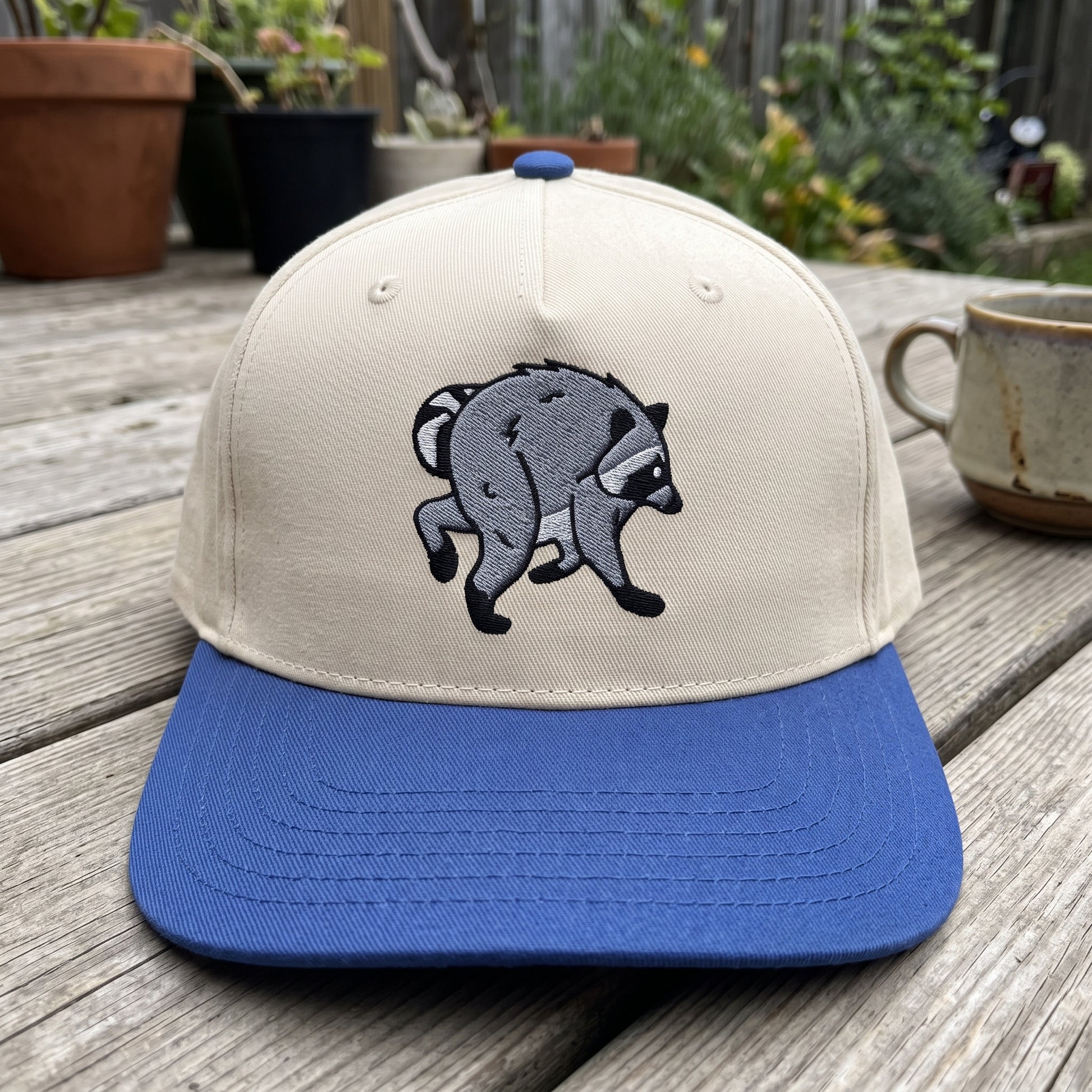Jimothy Raccoon embroidered hat in Royal/natural resting on a flat surface